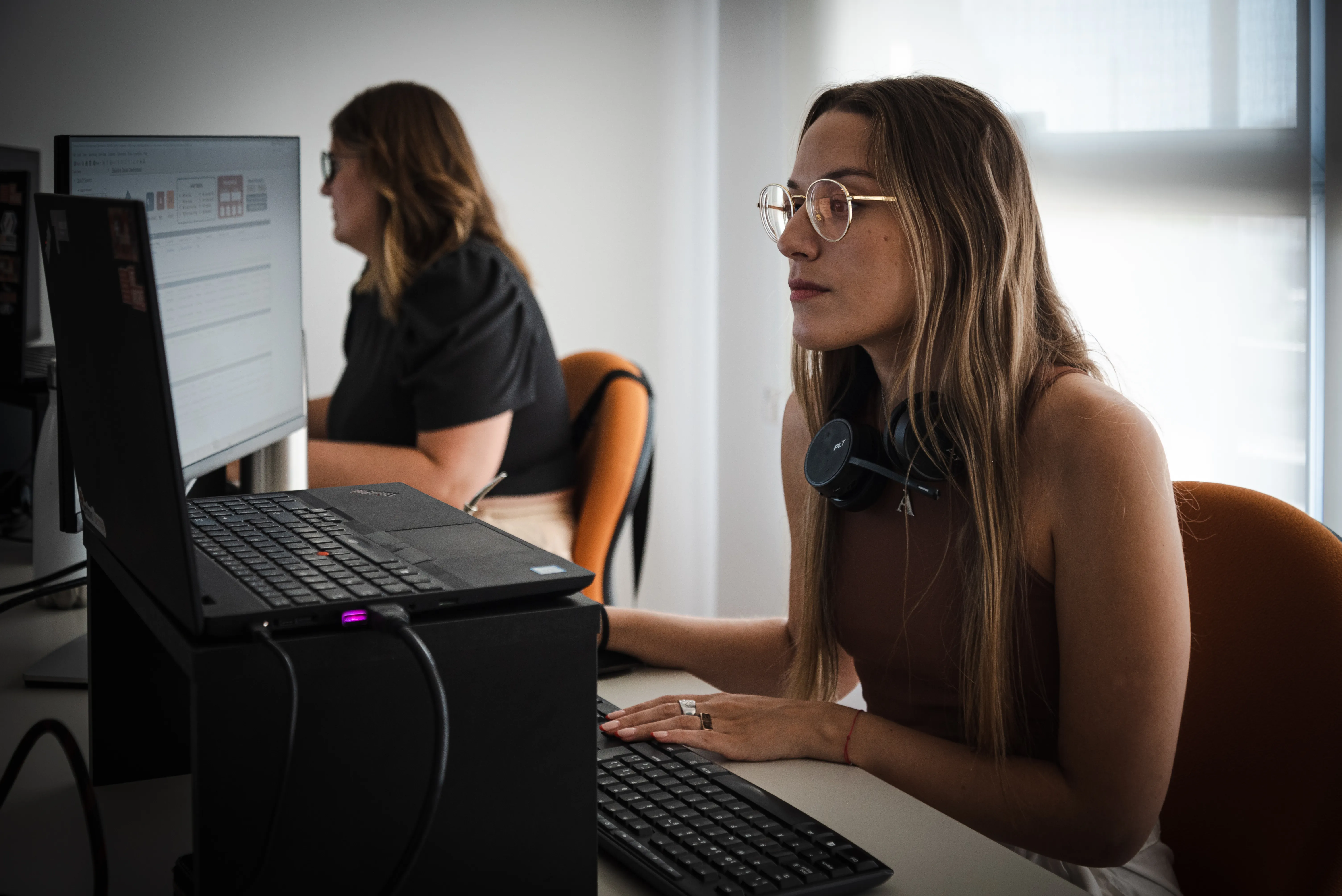 Two women are sitting at their desks, working on computers.
