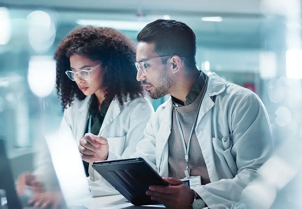 Researchers in lab coats analyzing data on a tablet and laptop in a modern laboratory.