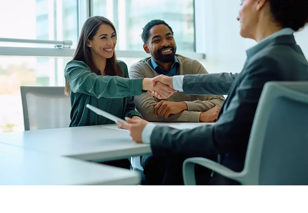 Smiling professionals in a business meeting, with two people shaking hands across the table.