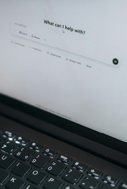 Laptop keyboard with a blurred screen showing an AI chat interface and the text “What can I help with?”