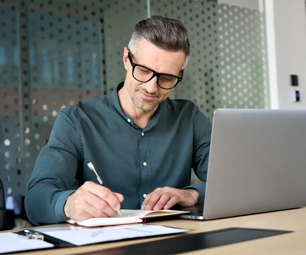 Professional taking notes while working on a laptop in a modern office setting.