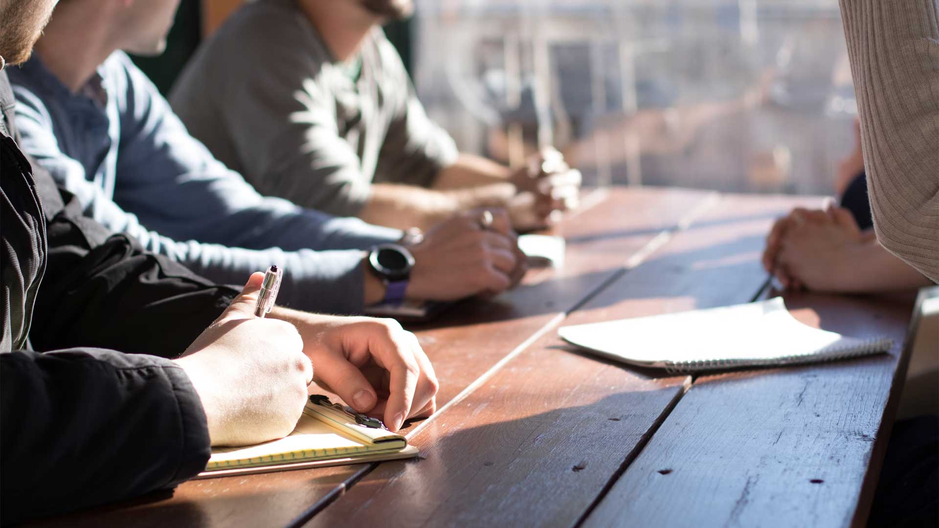 Several people seated at a table, engaged in discussion, while one individual is focused on writing